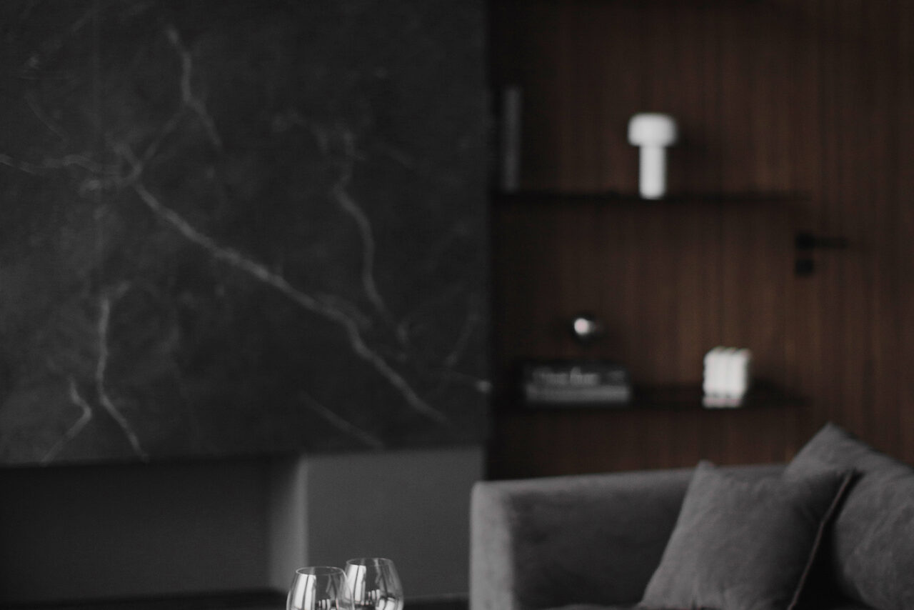 Арендовать Mimika(Мимика). Просторная фотостудия. Современная гостиная с темной мраморной стеной, коричневой деревянной панелью и полками. На фоне темный диван, на переднем плане стеклянные бокалы на столе. Освещение приглушенное, цвета преимущественно серые и коричневые.