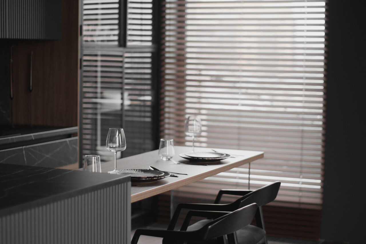 Арендовать Mimika(Мимика). Просторная фотостудия. Современная кухня с деревянным столом, сервированным на две персоны. Черные стулья, мраморная столешница, стеклянные бокалы. Большие окна с горизонтальными жалюзи, мягкое естественное освещение. Стены и мебель в нейтральных темных тонах.