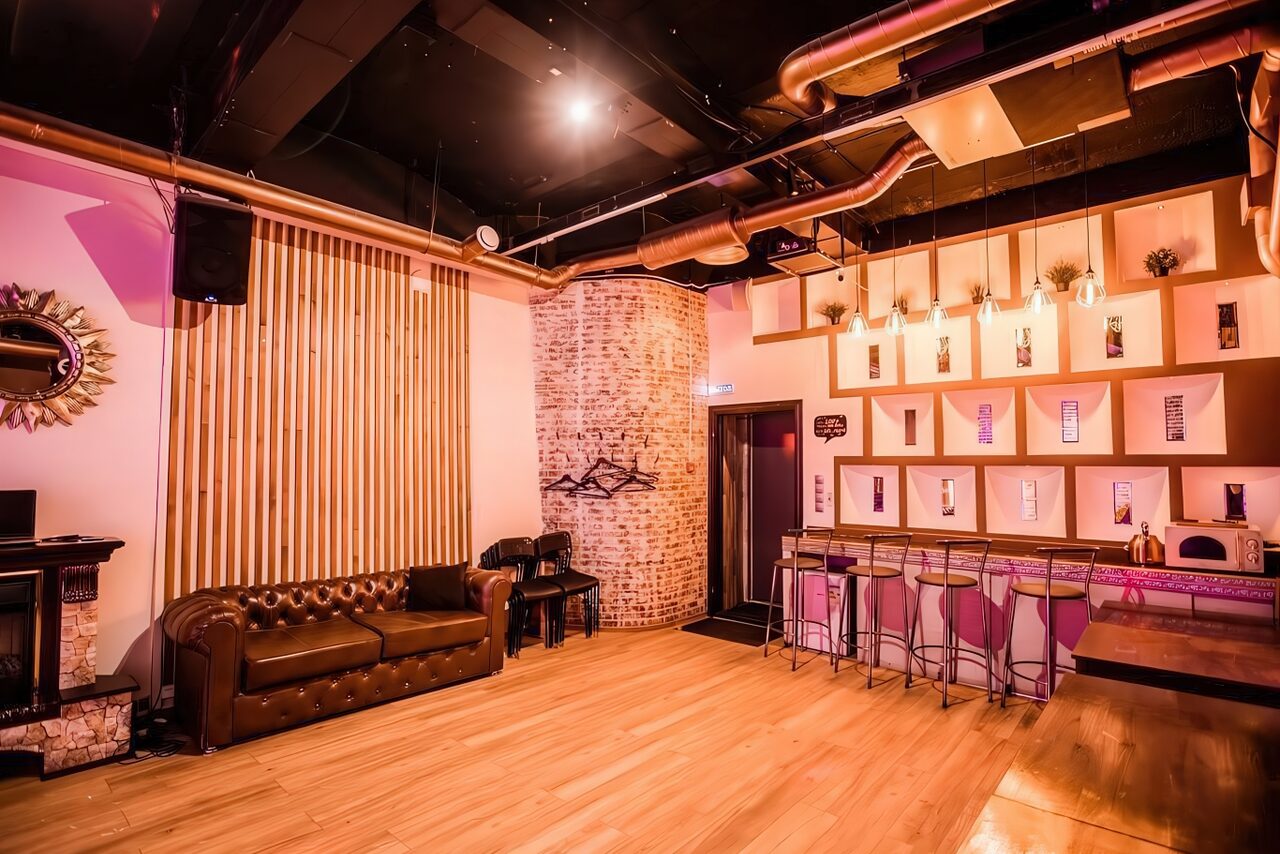 Арендовать LoftForParty. Бэкстейдж лофт . Лофт-пространство с барной зоной и кирпичной стеной. Диван и столы из темного дерева, барные стулья на высоких ножках. Пол из светлого дерева. Рейки на стене, декоративные лампы на потолке, открытые вентиляционные трубы. Теплое освещение.