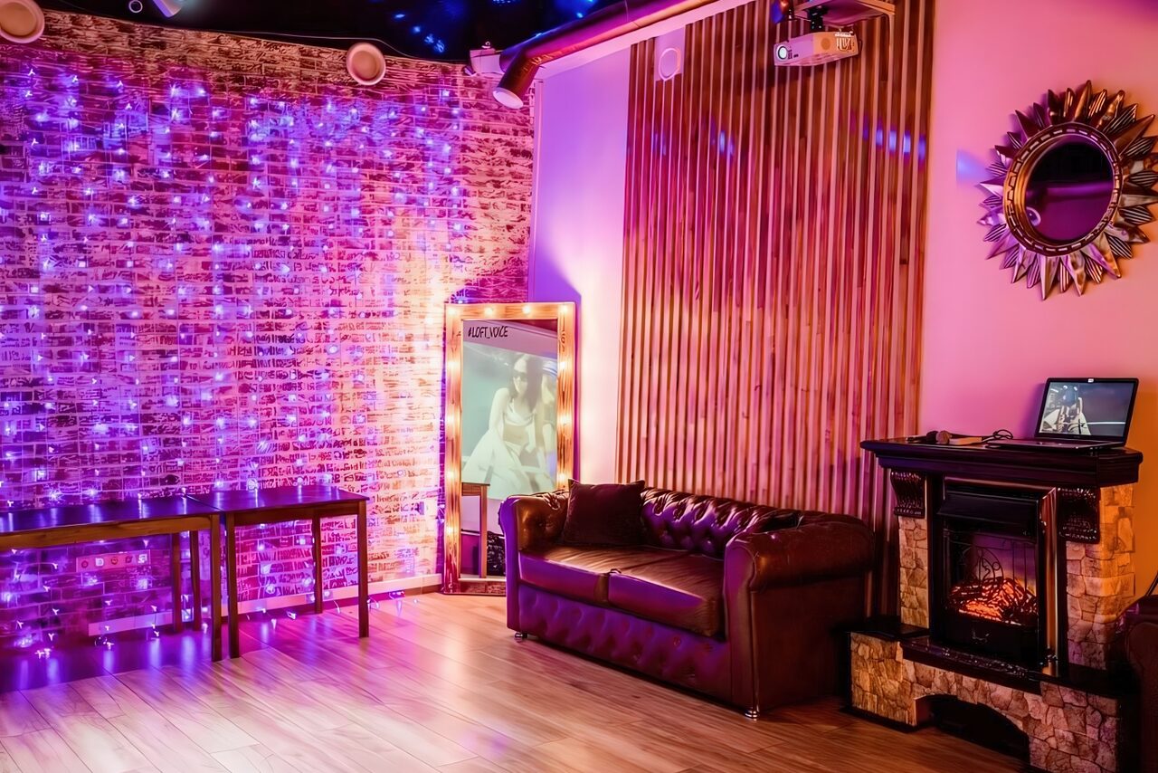 Арендовать LoftForParty. Бэкстейдж лофт . Лофт-пространство с кирпичной стеной, украшенной гирляндами фиолетового цвета. Коричневый кожаный диван, камин, зеркало в раме, пол из светлого дерева. Стена с деревянными панелями, стол и ноутбук. Круглое зеркало на стене.
