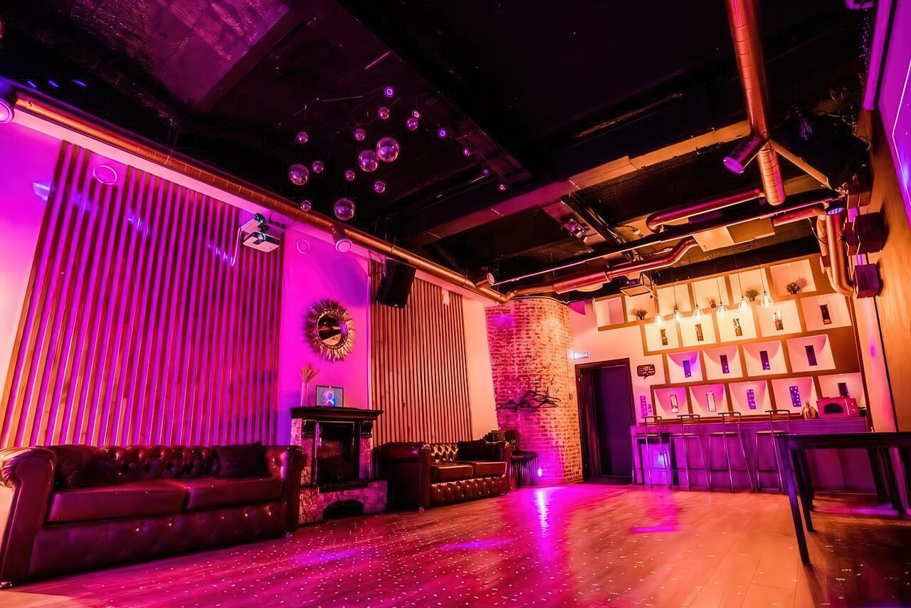 Арендовать LoftForParty. Бэкстейдж лофт . Клубное помещение с фиолетовым освещением, черным потолком и открытыми трубами. Два кожаных дивана у стен, барная стойка с высокими табуретами и полки. Деревянный пол, зеркальные шары на потолке, кирпичная стена, декоративные панели.