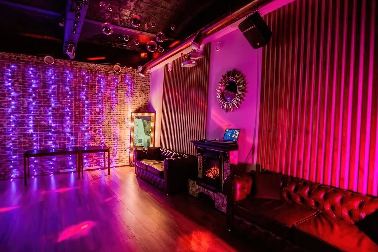 Арендовать LoftForParty. Бэкстейдж лофт . Лофт с кирпичными стенами и деревянным полом, оборудованный кожаными диванами. Освещение разноцветное, присутствуют неоновые гирлянды. На стене зеркало с декоративной рамой. Звуковое оборудование и ноутбук. Стены частично обшиты деревянными панелями.