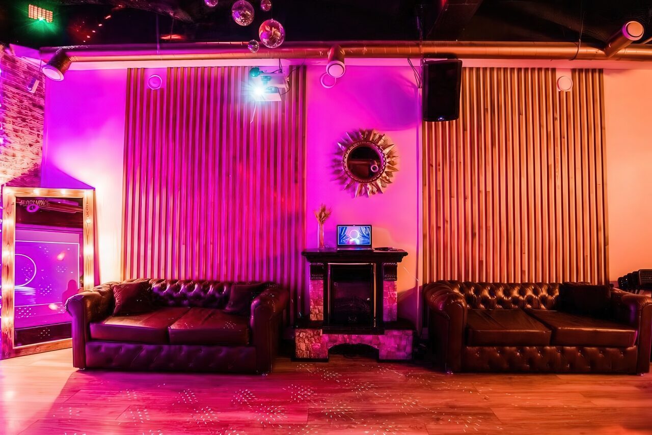 Арендовать LoftForParty. Бэкстейдж лофт . Лофт-пространство с кожаными диванами, прямоугольная планировка. Стена с деревянными панелями и зеркалом, камин с ноутбуком сверху. Яркое фиолетовое освещение, кирпичная кладка слева. Потолок с трубами и диско-шарами, светильники. Пол из светлого дерева.