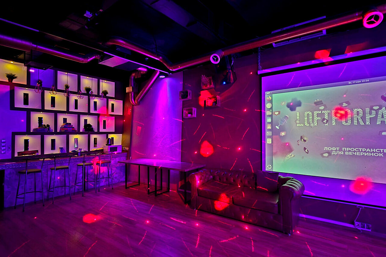 Арендовать LoftForParty. Бэкстейдж лофт . Тёмный лофт с неоновым освещением, барной стойкой и высокими стульями. На стене большой экран. Кожаный диван стоит у стены. Декоративные растения на полках. Потолочные трубы, деревянный пол. Надпись на экране: "Loft-пространство для вечеринок".