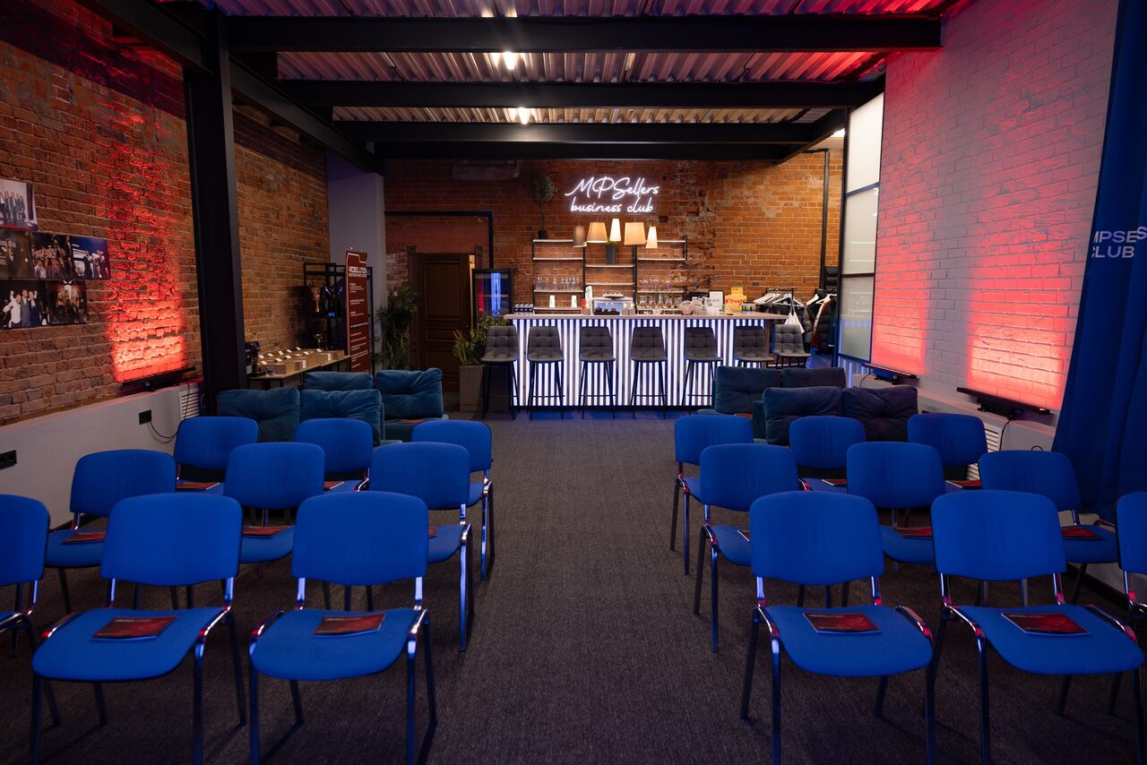 Арендовать MPSellers(МПСеллерс). Просторный зал для мероприятий. Пространство с кирпичными стенами, ряды синих стульев, барная стойка с неоновой надписью, сцена с микрофонами, мягкое боковое освещение, транспортная металлическая конструкция под потолком.