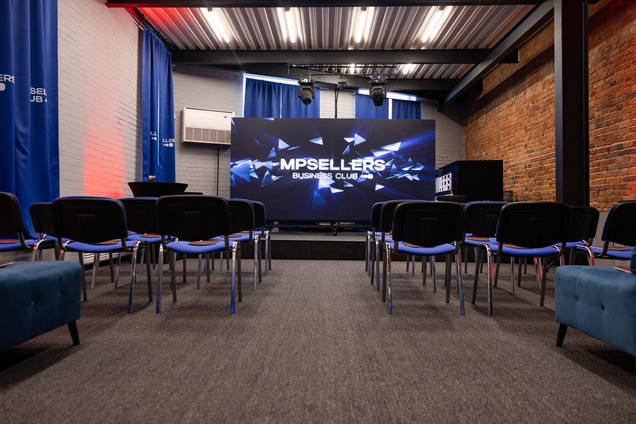 Арендовать MPSellers(МПСеллерс). Просторный зал для мероприятий. Аудитория с экраном, стулья в несколько рядов, серый ковролин, кирпичная стена справа, белая и серая кирпичная стена слева, синие шторы, подвесной потолок с освещением, кондиционер, надпись «MPSELLERS BUSINESS CLUB» на экране.