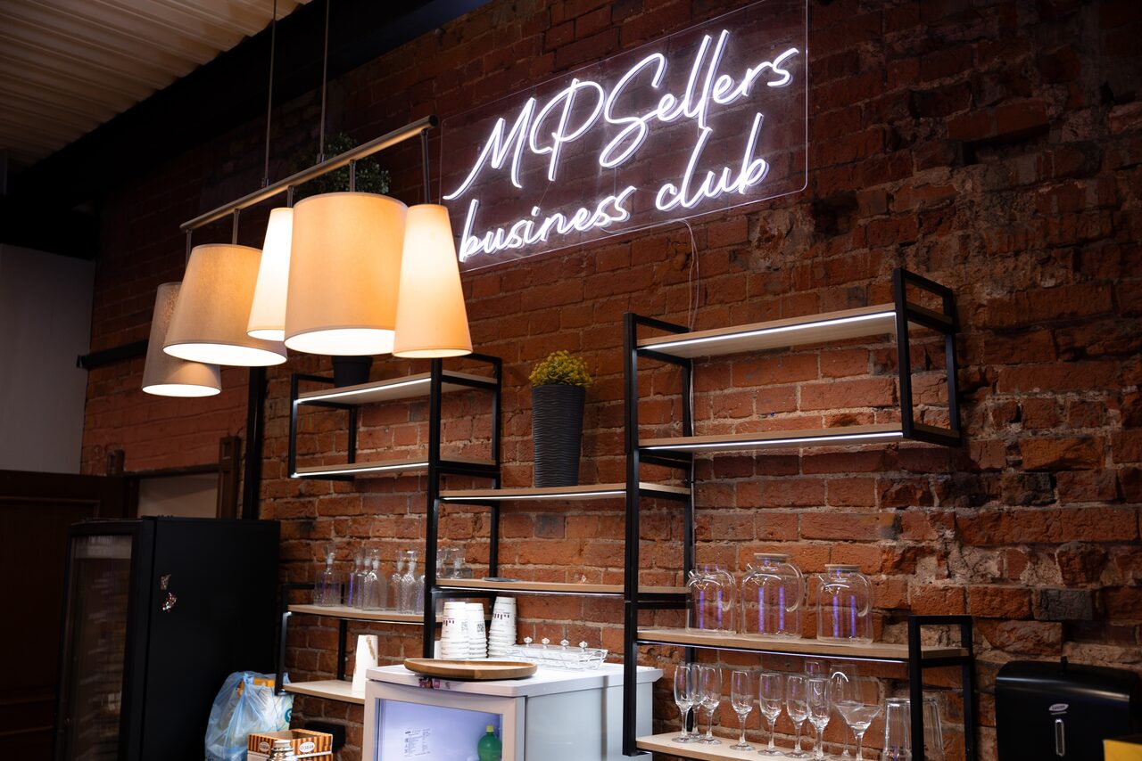Арендовать MPSellers(МПСеллерс). Просторный зал для мероприятий. Лофт-пространство с кирпичными стенами, стеллажи с посудой, подвесные лампы с абажурами, белый холодильник сбоку. Неоновая надпись "MPSellers business club". Декоративный горшок с растением на стеллаже.