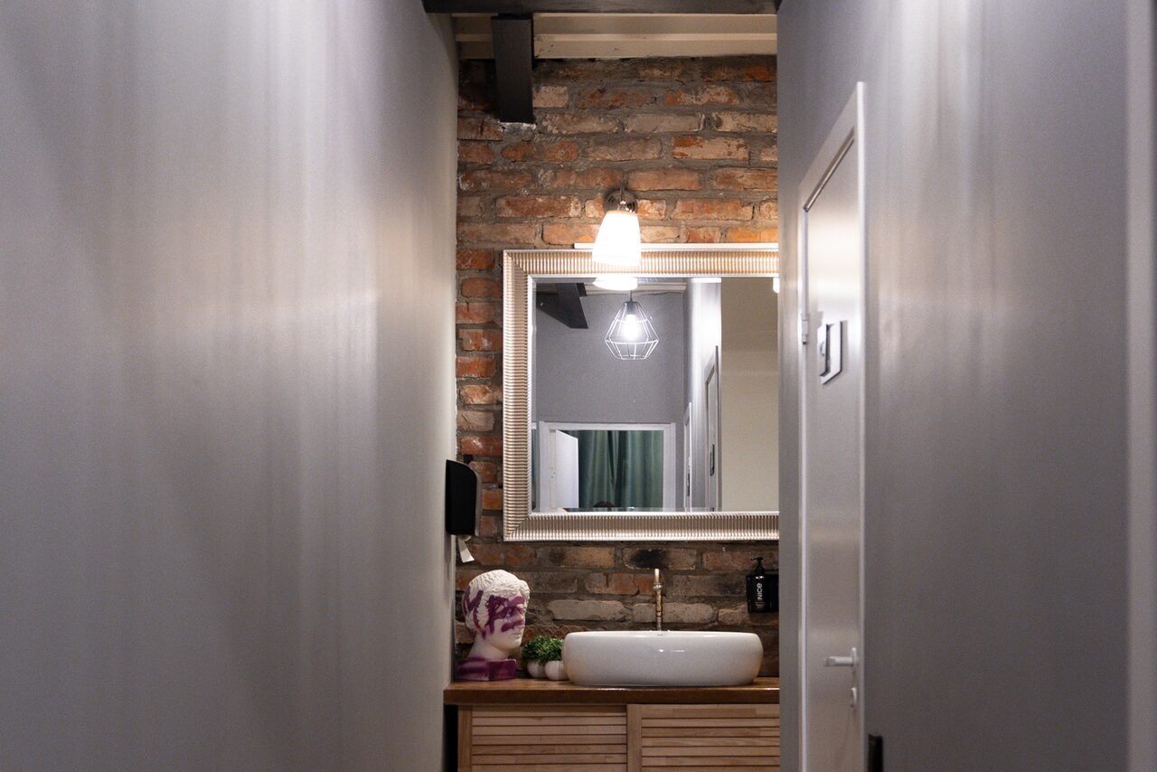 Арендовать MPSellers(МПСеллерс). Просторный зал для мероприятий. Узкий коридор с кирпичной стеной, белыми боковыми стенами, подвесным светильником, зеркалом в раме над раковиной на деревянной тумбе. Иллюзорное отражение в зеркале, сбоку диспенсер для мыла, сверху настенный светильник.