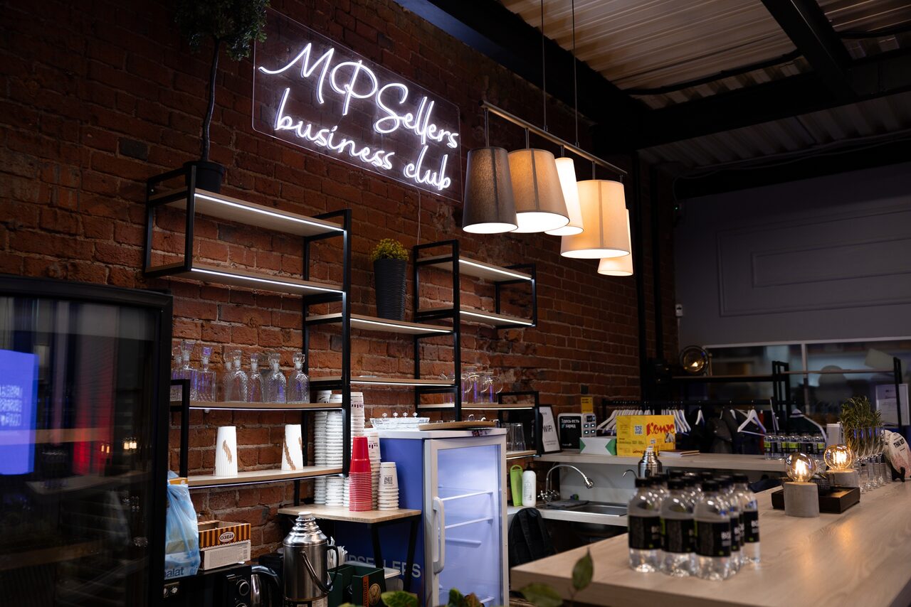 Арендовать MPSellers(МПСеллерс). Просторный зал для мероприятий. Кафе с кирпичными стенами и деревянной стойкой. Неоновая вывеска "MP Sellers business club". Открытые полки с посудой, ламинированная стойка, подвесные светильники, холодильник, белая и серебристая посуда, бутылки с водой на стойке.