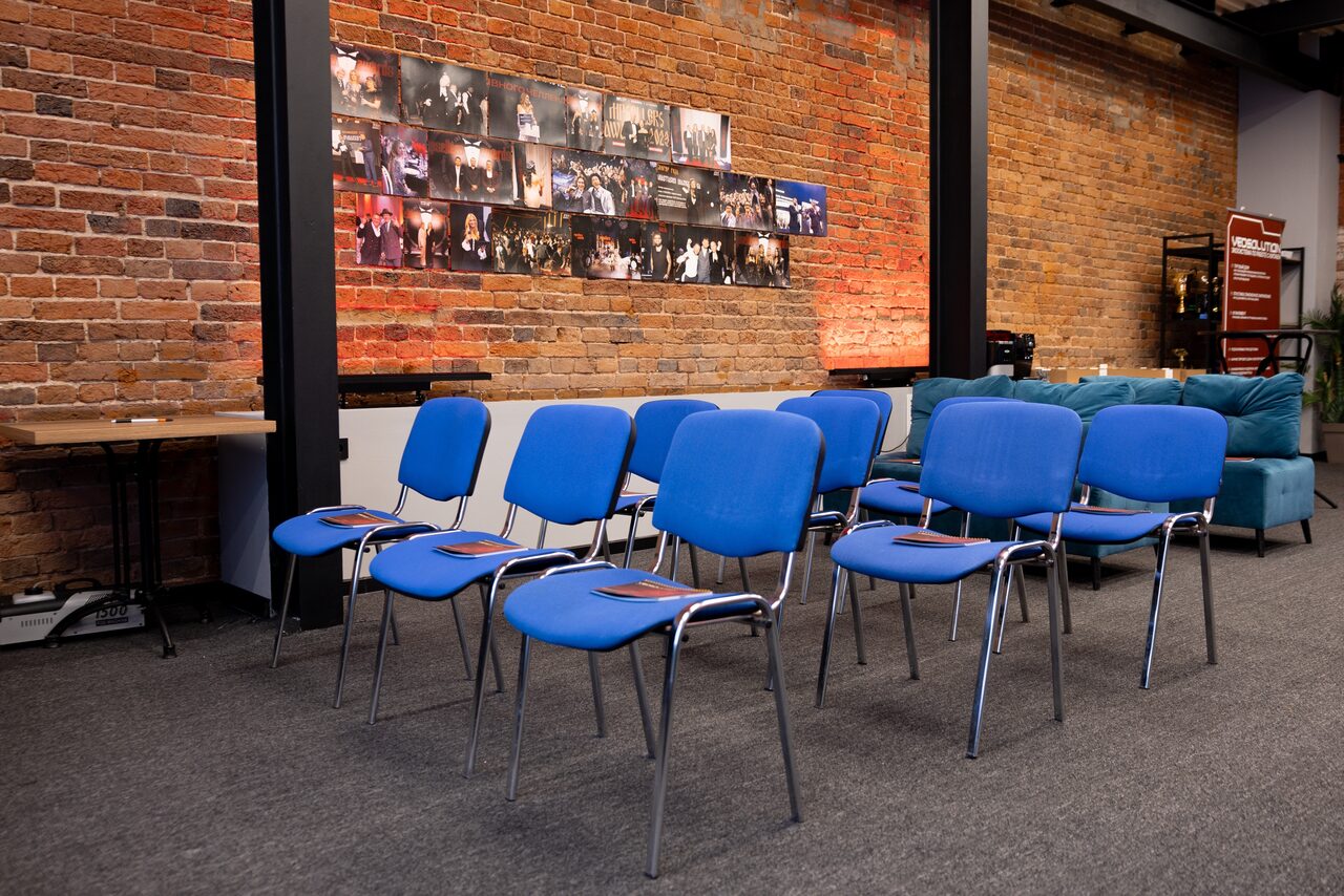 Арендовать MPSellers(МПСеллерс). Просторный зал для мероприятий. Ряд синих стульев на сером ковровом покрытии, кирпичная стена с фотоколлажем, деревянный стол слева, мягкие синие кресла справа, освещение с потолка, металлические колонны.