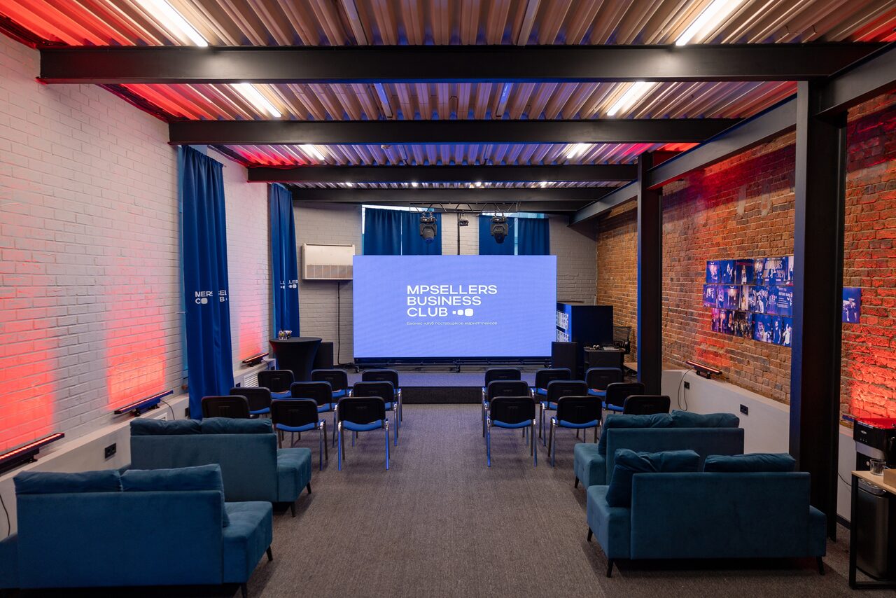 Арендовать MPSellers(МПСеллерс). Просторный зал для мероприятий. Пространство с проектором и экраном, ряды стульев перед ним, кирпичная и светлая стена, металлический потолок с черными балками, синие шторы, диваны. Освещение разноцветное, с акцентом на красный и синий. Текст на экране: "MPSELLERS BUSINESS CLUB".