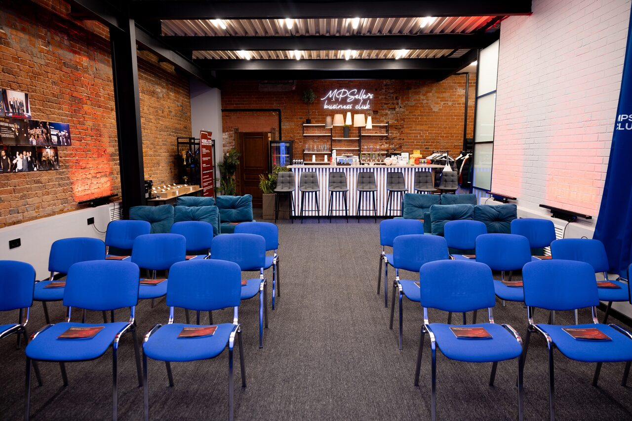 Арендовать MPSellers(МПСеллерс). Просторный зал для мероприятий. Пространство в стиле лофт с кирпичными стенами, металлическими балками и серым ковровым покрытием. Расставлены синие стулья в два ряда, диваны у стен, на заднем плане барная стойка. Светодиодная вывеска, яркое освещение.