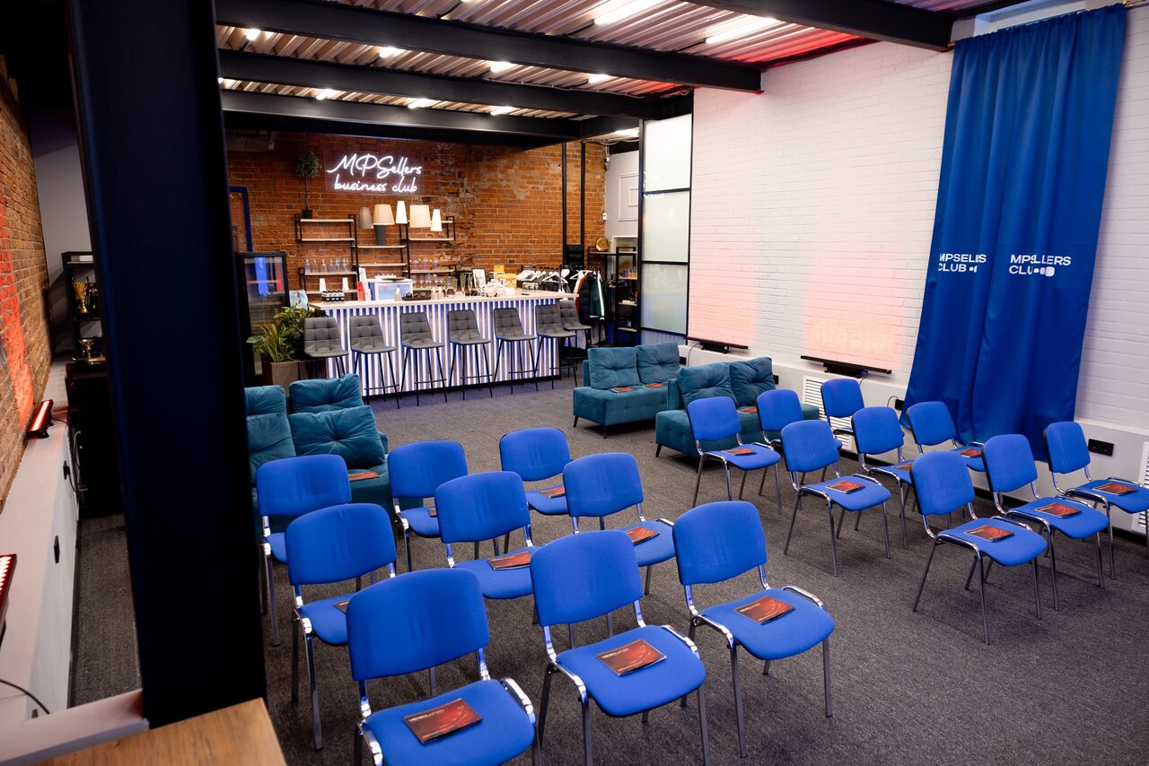 Арендовать MPSellers(МПСеллерс). Просторный зал для мероприятий. Многофункциональное пространство с синими стульями в ряд, синими диванами, кирпичной стеной и барной стойкой с белыми лампами. Стена украшена неоновой надписью. Синий занавес. Серый ковер. Стиль лофт.
