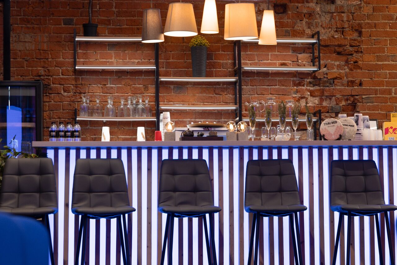 Арендовать MPSellers(МПСеллерс). Просторный зал для мероприятий. Барная стойка с высокими черными стульями, кирпичная стена, металлические полки с бутылками и бокалами, лампы разных форм, синий и белый свет. На стойке стеклянные и пластиковые емкости, есть цветочный горшок и различные упаковки.