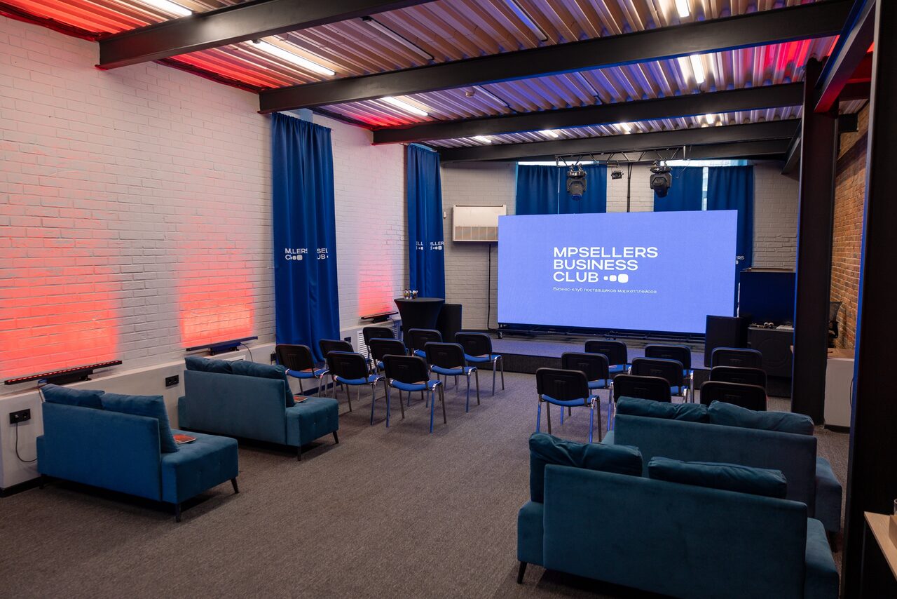 Арендовать MPSellers(МПСеллерс). Просторный зал для мероприятий. Просторное помещение с рядами черных стульев, несколькими голубыми диванами и проекционным экраном. Белые кирпичные стены с красной подсветкой, синие занавески, серый ковер. Металлический потолок с подвесными светильниками и кондиционер на стене. Текст на экране: "MPSELLERS BUSINESS CLUB".