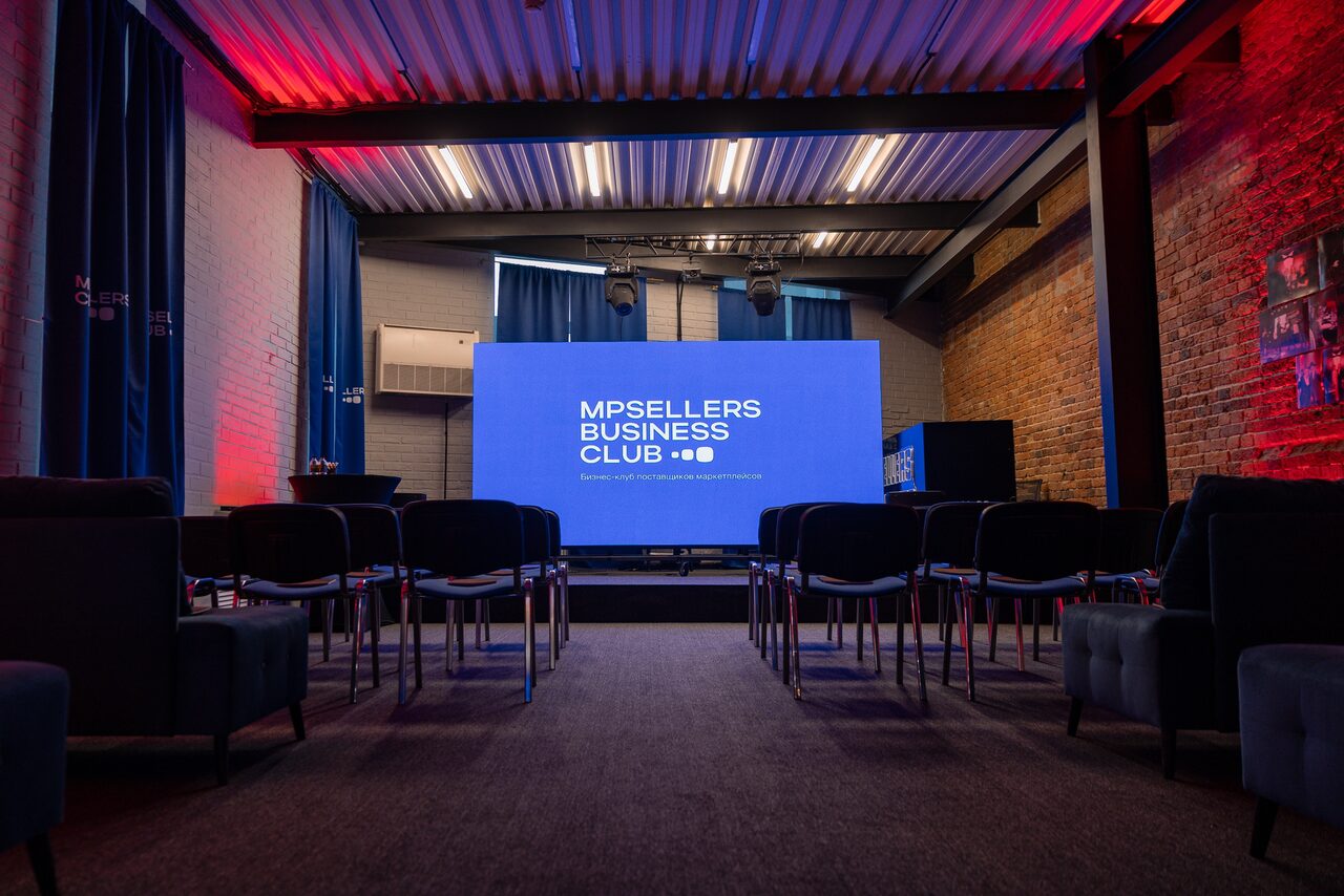 Арендовать MPSellers(МПСеллерс). Просторный зал для мероприятий. Небольшой зал с высоким потолком и полом, обитым ковролином. Ряды стульев перед большим экраном с надписью "MPSELLERS BUSINESS CLUB". Открытые кирпичные стены, синие шторы, подвесное освещение, металлические элементы на потолке.