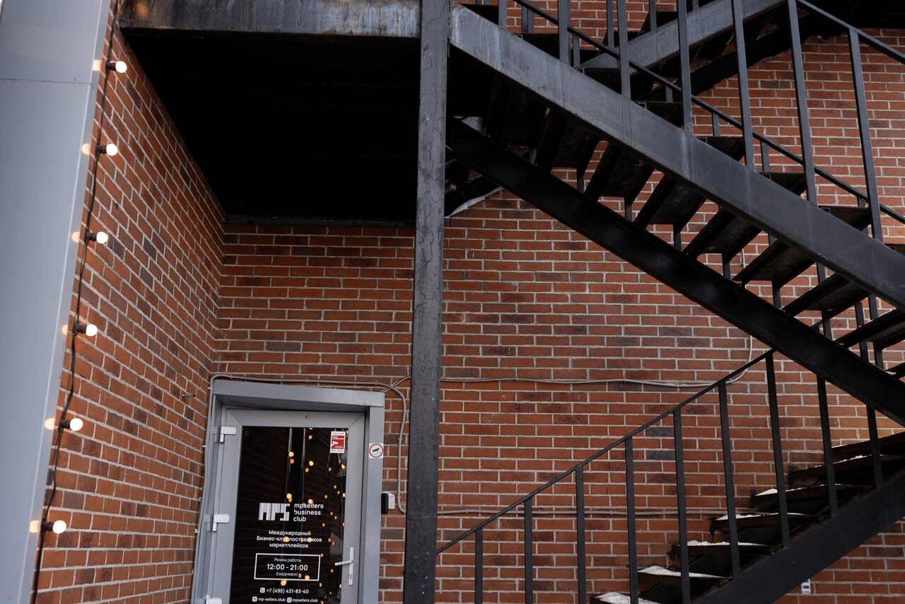 Арендовать MPSellers(МПСеллерс). Просторный зал для мероприятий. Красно-кирпичная стена с черной металлической лестницей. Входная дверь со стеклянной вставкой и изображением логотипа и информации о часах работы. Освещение вдоль стены представлено гирляндой из круглых лампочек.