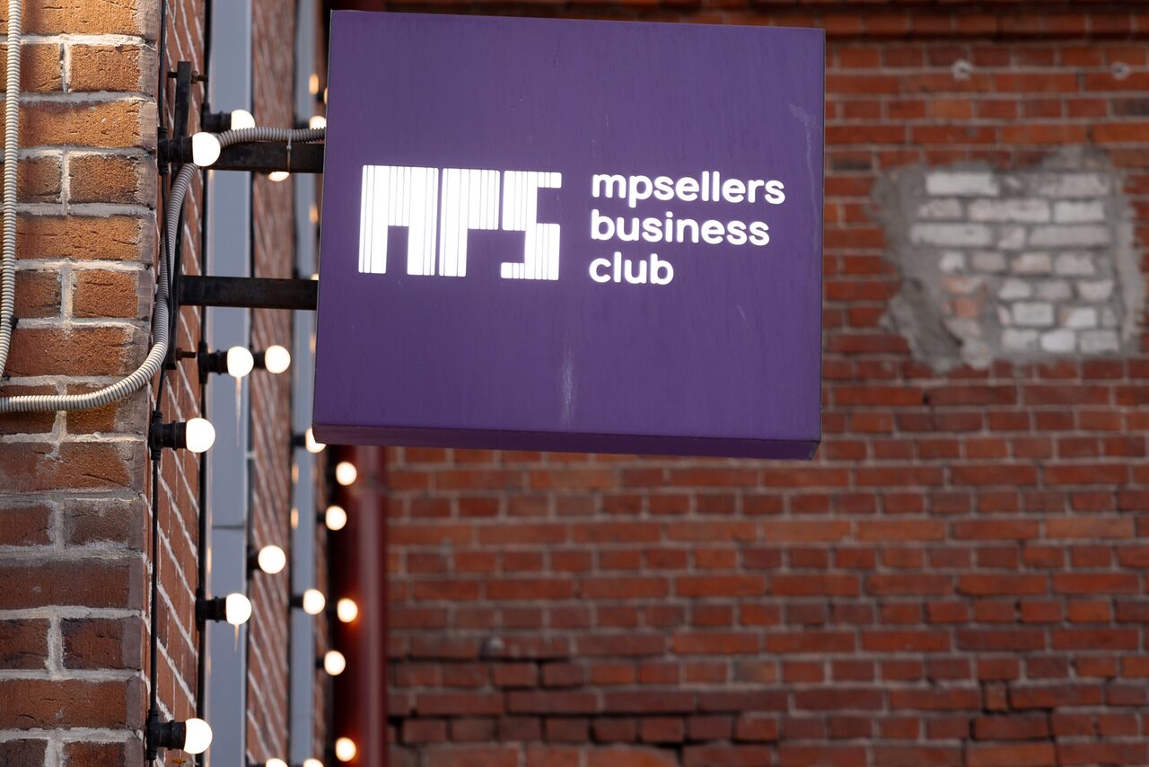 Арендовать MPSellers(МПСеллерс). Просторный зал для мероприятий. Вывеска с надписью "mpsellers business club" на фиолетовом фоне, закреплена на кирпичной стене. По левую сторону видна гирлянда с лампами. Стена частично побелена в одном месте.