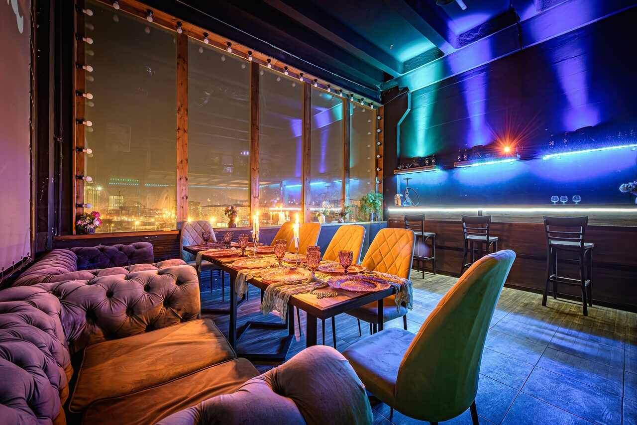 Арендовать Лофт ZUM(Зум). Пространство с мягкими диванами и стульями вокруг обеденного стола, сервированного посудой. Большие окна со шторками, светодиодная подсветка на стенах. Потолок тёмный, пол плиточный. Декоративные огни вдоль стены, барная стойка со стульями.