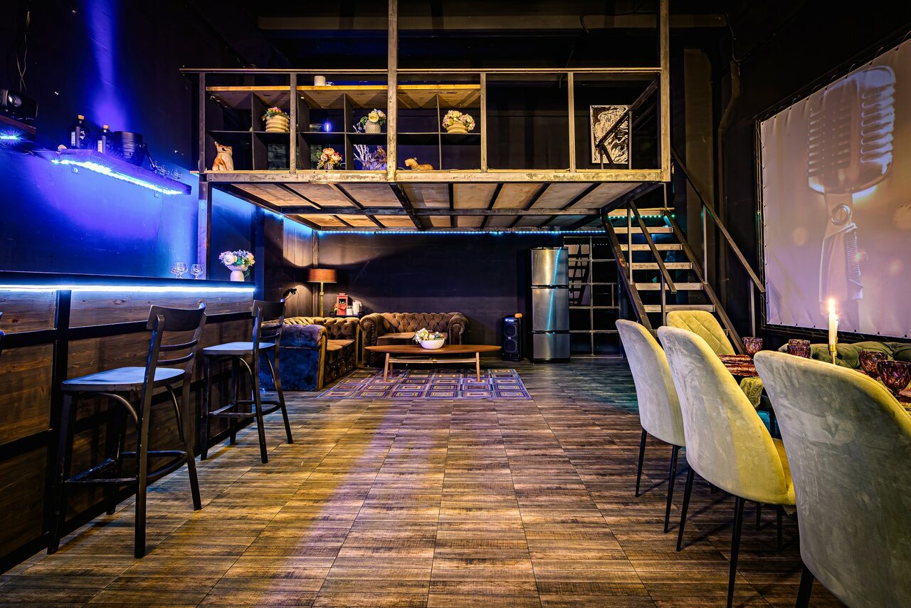 Арендовать Лофт ZUM(Зум). Лофт-пространство с мезонином и деревянной лестницей, креслами и высокими стульями, деревянный пол, неоновая подсветка, темные стены, зона отдыха с диваном, журнальный столик, холодильник, настольная лампа, декоративные элементы на втором уровне.