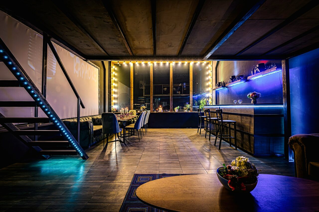 Арендовать Лофт ZUM(Зум). Лофт с деревянным полом и открытыми потолочными балками. Лестница с подсветкой. Большие окна с гирляндой ламп. Барная стойка с подсветкой, высоким барным стульями. Диван вдоль стены и стол с цветочным декором. Тёмные и синие цвета.