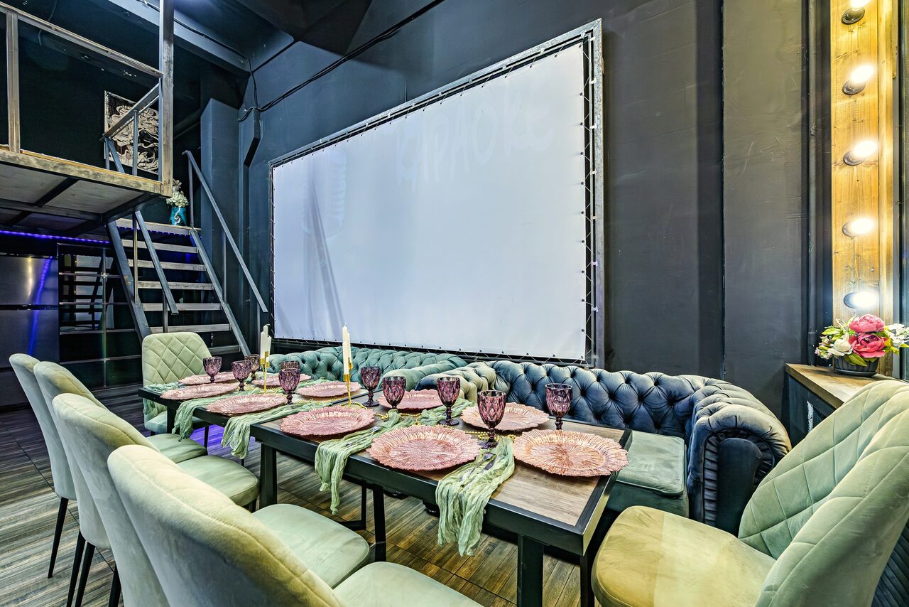 Арендовать Лофт ZUM(Зум). Современное пространство с деревянным полом и черными стенами. Обеденная зона: длинный стол с розовыми плетеными подставками, сервирован фужерами и подсвечниками. Мягкие кресла, диван. Большой экран. Лестница на второй уровень. Настенные лампочки.