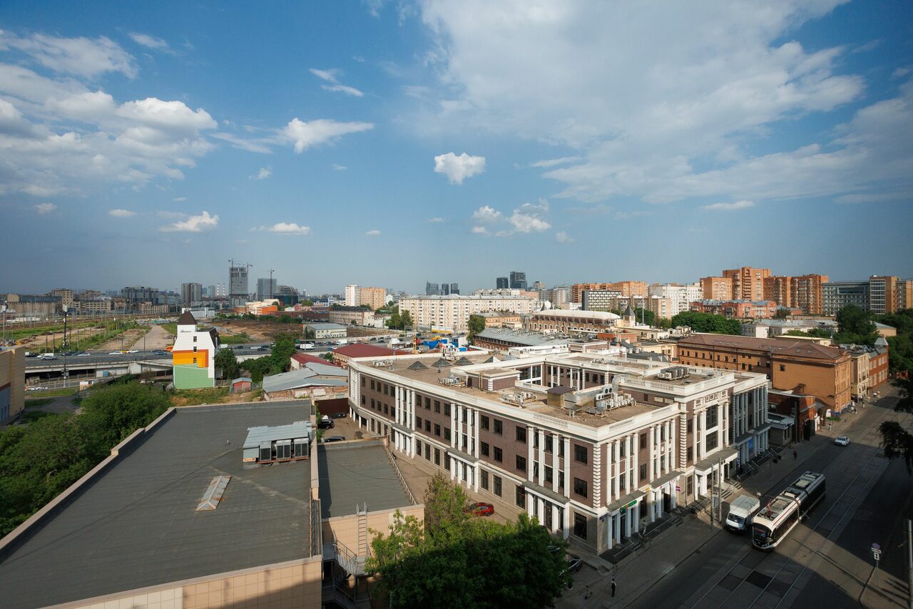 Арендовать Prostranstvo (Пространство). Лофт ZUM (Зум). Городской пейзаж с видом на улицы и крыши зданий. Видны многоэтажные здания, дороги и деревья. Яркий солнечный день с голубым небом и облаками.
