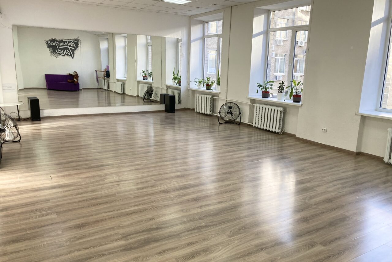 Арендовать Территория Танца. Просторное помещение с деревянным полом и большими окнами. На одной стене большое зеркало. Вдоль окон размещены растения, радиаторы и вентиляторы. В углу фиолетовый диван. На стене текст. Светлые стены и потолок.