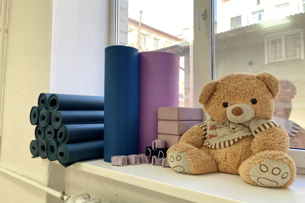 Арендовать Территория Танца. Подоконник с йога-ковриками синего и фиолетового цвета, мягкими блоками и ремнями для йоги, плюшевый медведь в полосатой кофте. Белая рама окна, вид на городские здания. Светлый интерьер.
