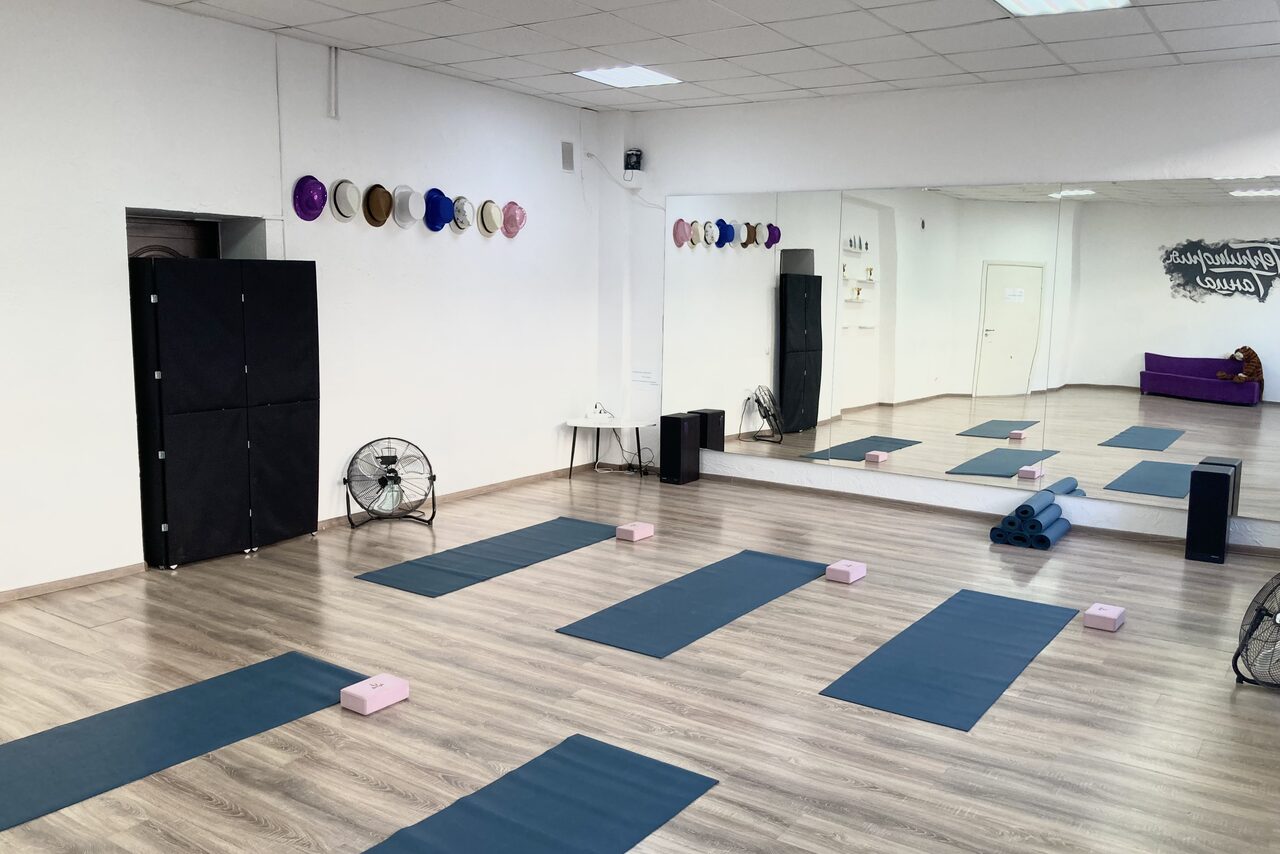 Арендовать Территория Танца. Просторный светлый зал с деревянным полом, большими зеркалами на стене и высоким потолком. Организованы коврики и блоки для йоги. На стене висят барабаны в чехлах. Присутствуют вентиляторы и черные колонки. Белые стены, дверной проем.