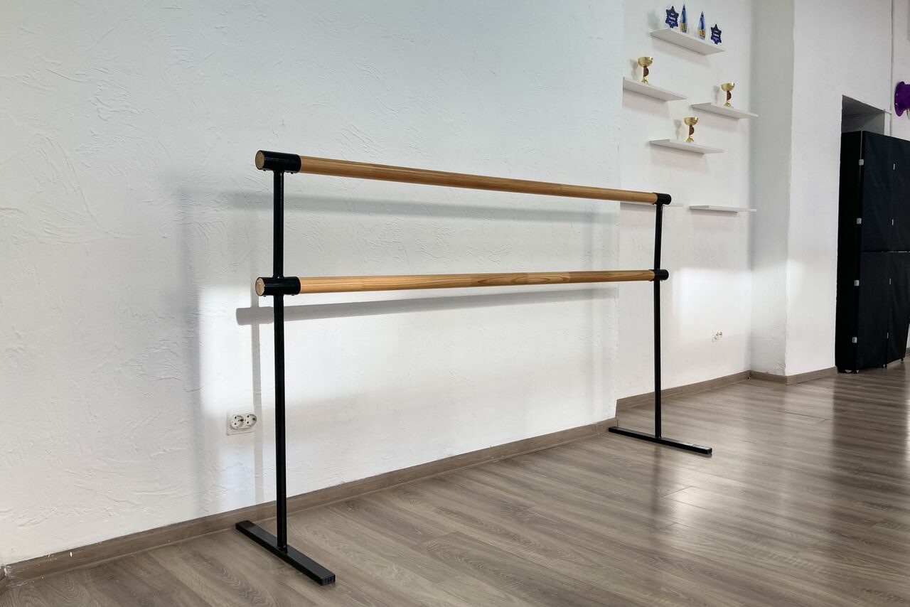 Арендовать Территория Танца. Зал с балетным станком на светло-коричневом ламинате. Белая стена с текстурой и открытыми полками с кубками. Чёрная мебель или оборудование у стены. Естественное освещение. Электрическая розетка на стене.