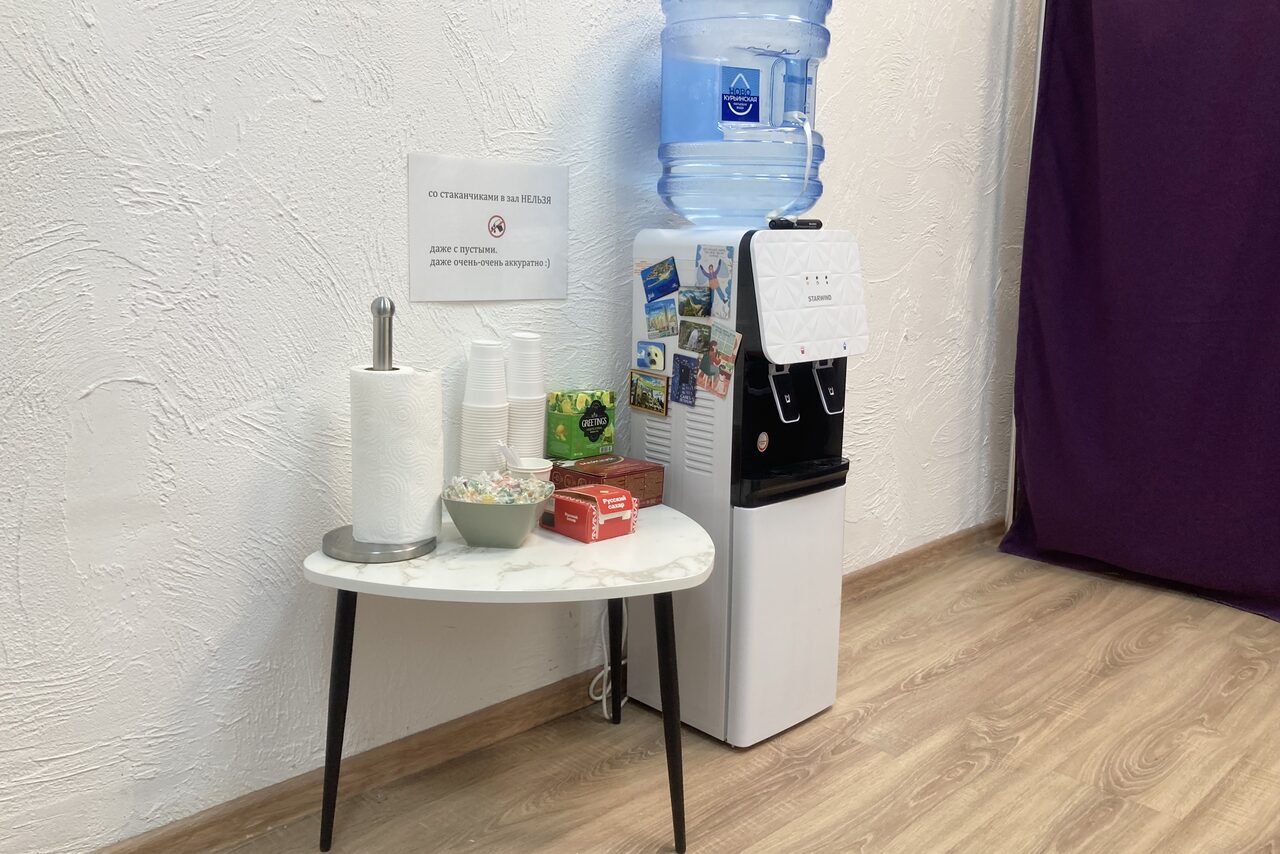 Арендовать Территория Танца. Стол с бумажными полотенцами, пластиковыми стаканчиками и леденцами рядом с кулером для воды. Белая стена с текстурой, на которой висит лист с текстом и изображениями. Напольный ламинат и фиолетовая штора на заднем плане.