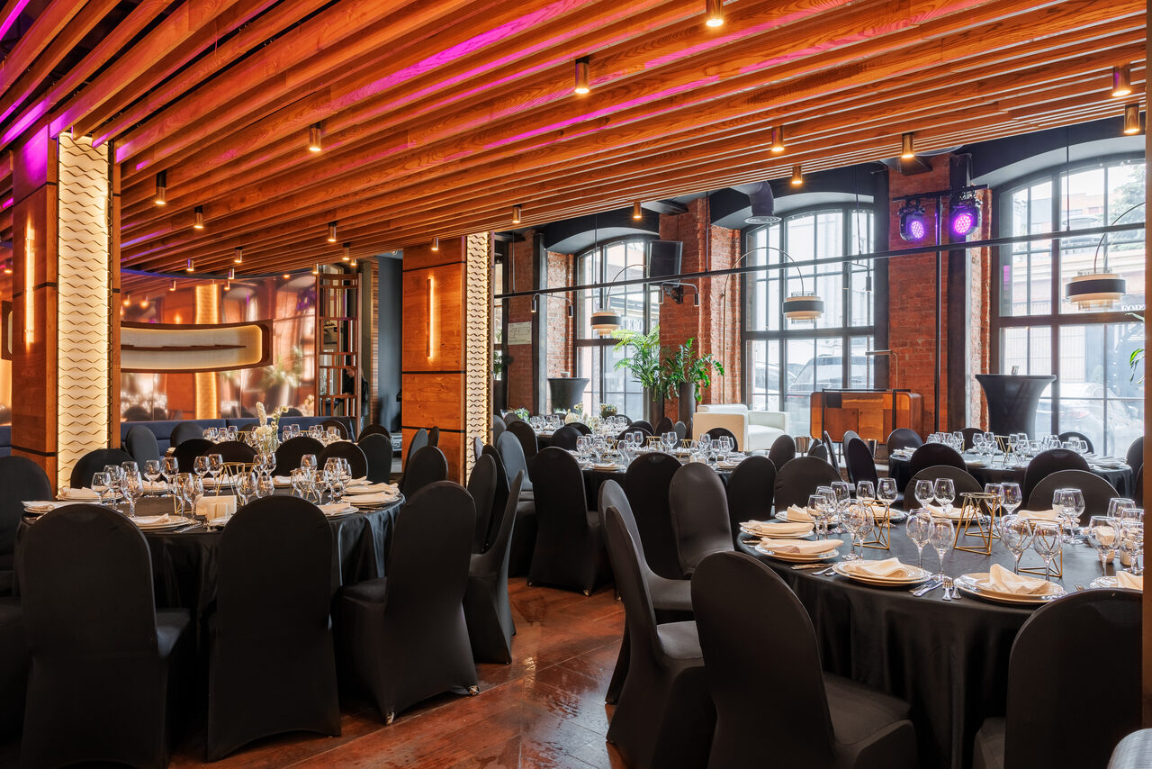 Арендовать Zafferano Loft(Зафферано лофт). Пространство с деревянным потолком и кирпичными стенами, круглыми столами, сервированными стеклянной и столовой посудой, черными чехлами на стульях. Большие окна, точечное освещение, подвесные светильники, зеленые растения, полки с книгами.