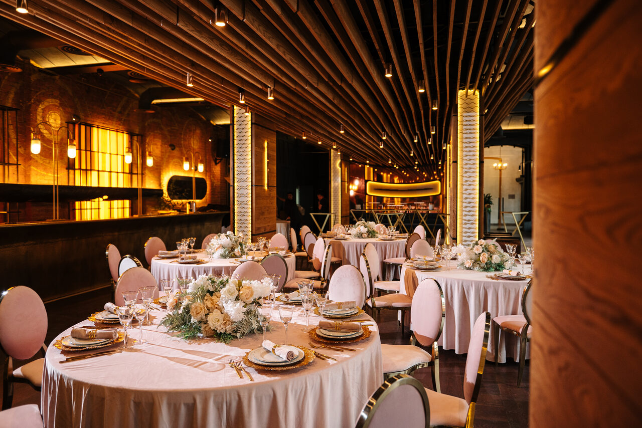 Арендовать Zafferano Loft(Зафферано лофт). Банкетный зал с круглой расстановкой столов, покрытых белыми скатертями и украшенных цветочными композициями. Розовые мягкие стулья с металлическими ножками. Потолок с деревянными балками и точечными светильниками. Стены кирпичные, теплое освещение.