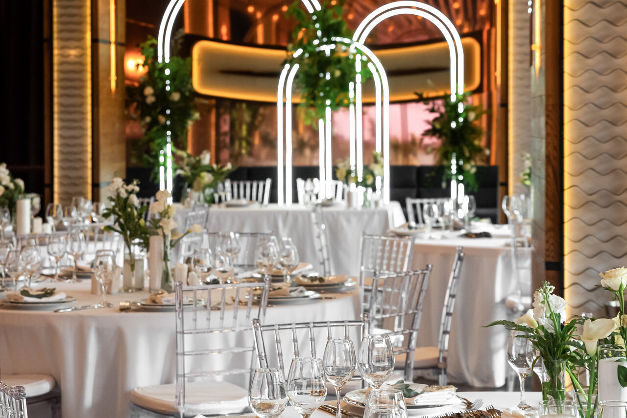 Арендовать Zafferano Loft(Зафферано лофт). Банкетный зал с круглыми столами, накрытыми белыми скатертями, и прозрачными стульями. На столах бокалы и белые цветы. Арки с неоновыми огнями и зеленью. Лёгкое декоративное освещение, стены с золотыми и белыми элементами.