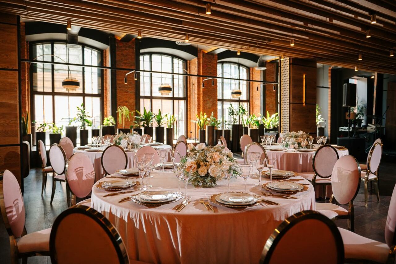 Арендовать Zafferano Loft(Зафферано лофт). Большой зал с круглыми столами, покрытыми белыми скатертями и сервированными тарелками и бокалами. Овальные стулья с белыми спинками. Высокие окна с черными рамами. Растения на подоконниках. Потолок с деревянными панелями и подвесными светильниками.