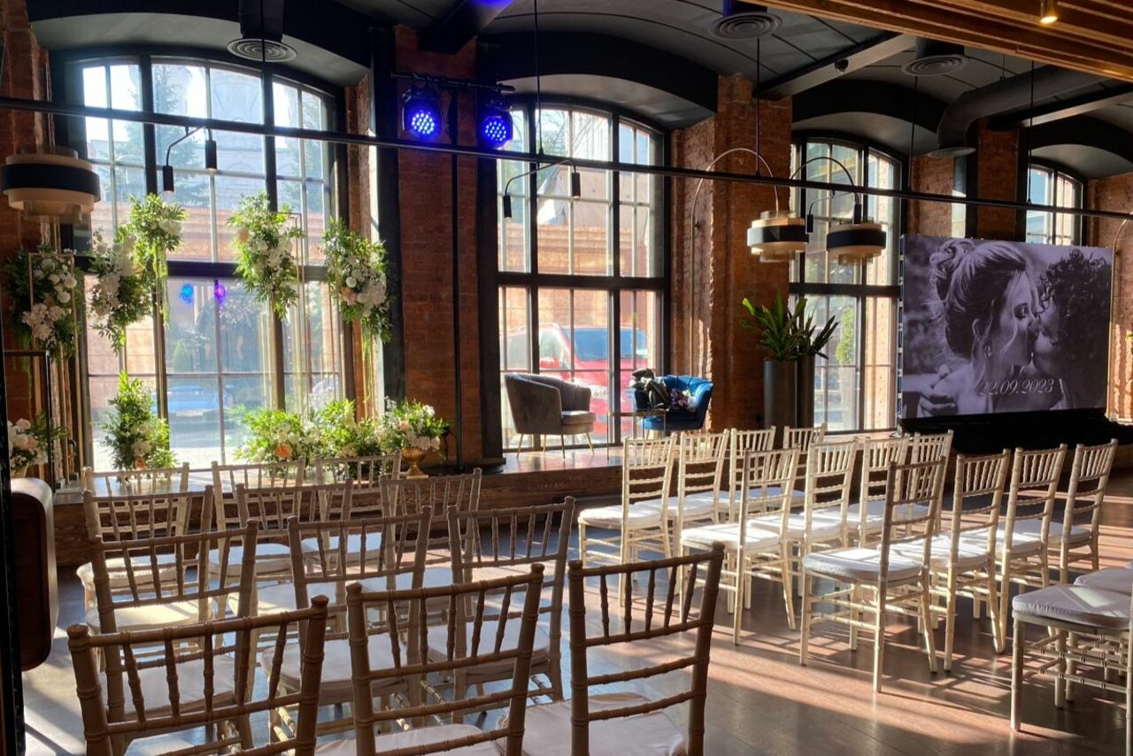 Арендовать Zafferano Loft(Зафферано лофт). Светлое пространство с кирпичными стенами и большими арочными окнами. Ряды деревянных стульев лицом к экрану с изображением. Подвесные светильники и декоративные растения. Потолок с деревянными балками, пол темный.