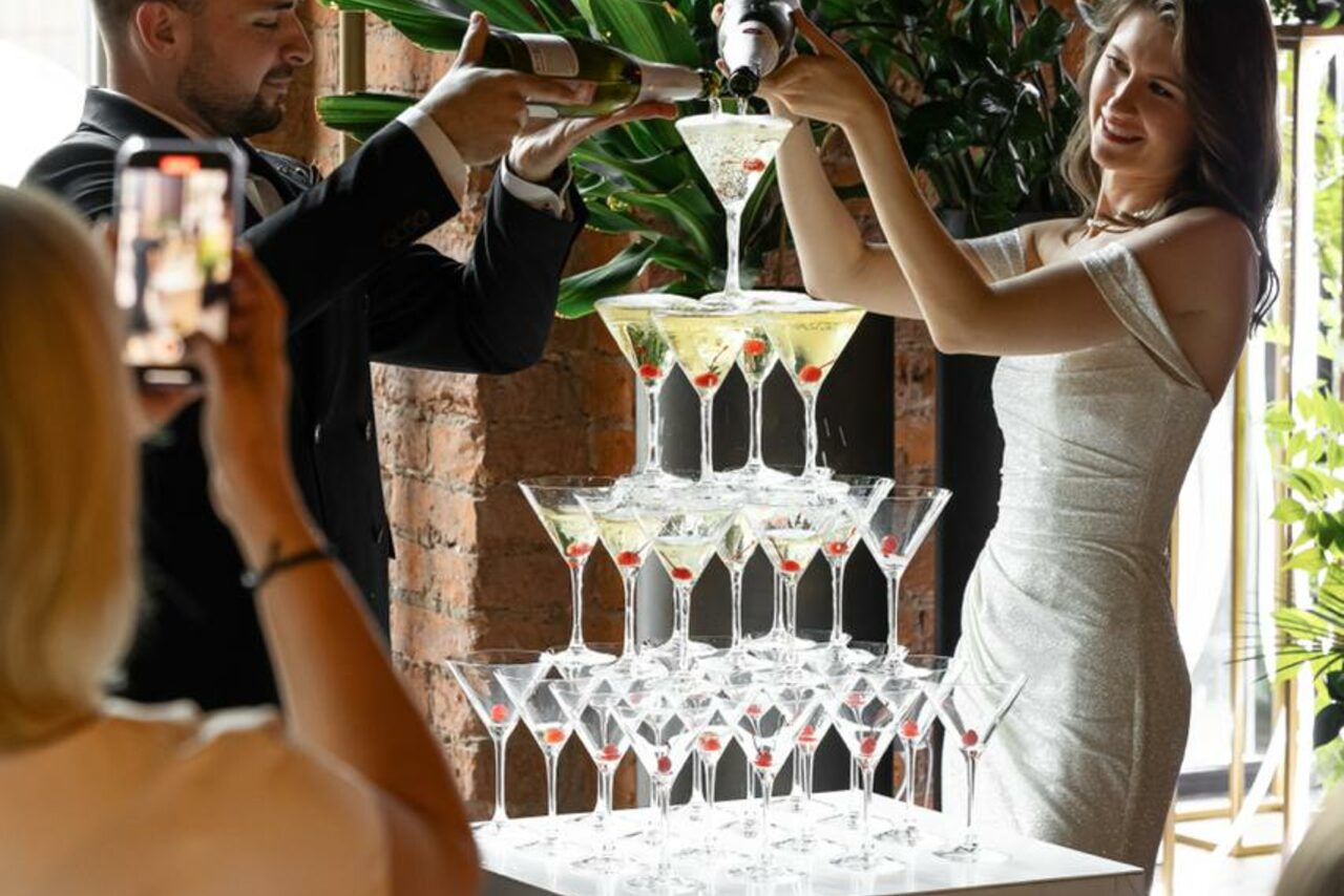 Арендовать Zafferano Loft(Зафферано лофт). Пирамидальная композиция из бокалов для коктейлей, наполненных шампанским, на белом столе. Люди в вечерних нарядах наливают напитки. Задний план с кирпичной стеной и крупными зелеными растениями. Яркое освещение.