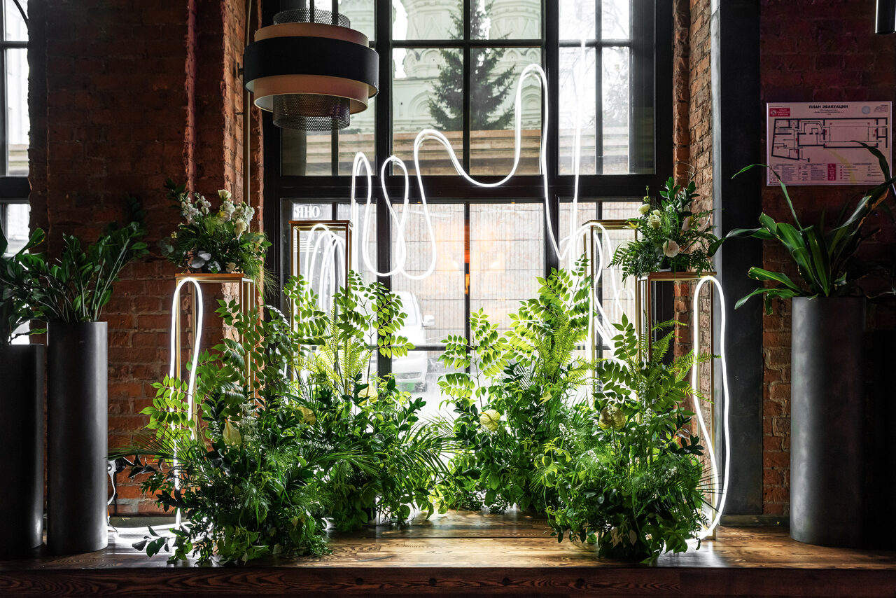 Арендовать Zafferano Loft(Зафферано лофт). Лофт-пространство с кирпичными стенами и большими окнами. На деревянной поверхности располагаются растения в высоких металлических кашпо. Светодиодные лампы изогнутой формы. Над столешницей подвесной светильник. На стене схема помещения.