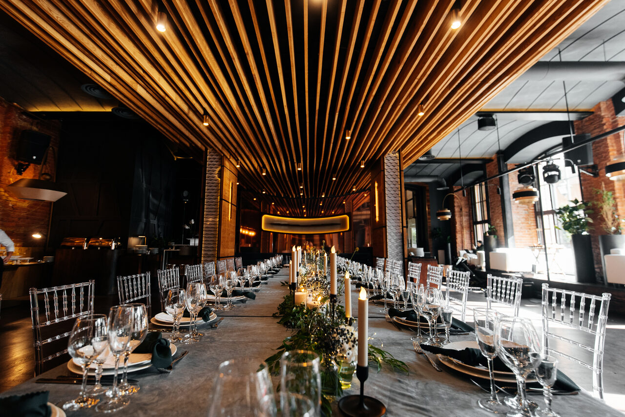 Арендовать Zafferano Loft(Зафферано лофт). Пространство для мероприятий с длинным обеденным столом, сервированным стеклянной посудой и приборами. Стулья прозрачные, потолок с деревянными панелями и встроенным освещением. Стены из кирпича, большие окна с черными рамами. Декор из зелени и свечей.