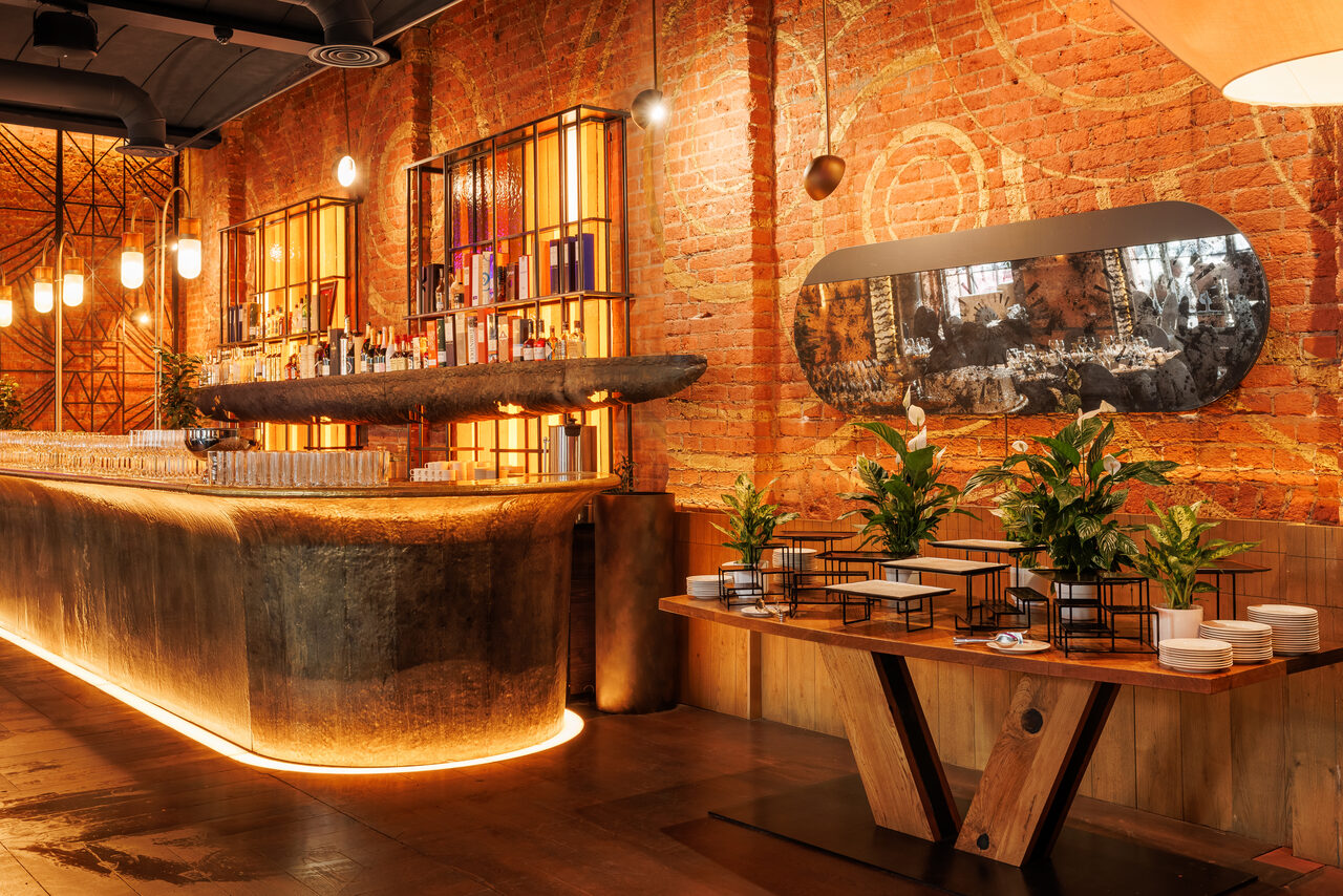 Арендовать Zafferano Loft(Зафферано лофт). Пространство бара с кирпичными стенами, деревянной столешницей и металлическими элементами. Зона стойки оформлена подсветкой. Размещены полки с бутылками. Растения в горшках на столе. Яркое верхнее освещение. Настенное зеркало овальной формы.