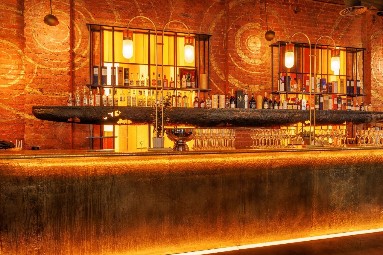 Арендовать Zafferano Loft(Зафферано лофт). Барное пространство с длинной деревянной стойкой, украшенной светодиодной подсветкой. Задняя стена из кирпича с круговыми узорами. На полках бутылки алкоголя. Подвесные светильники с мягким светом. Прозрачные бокалы на стойке.