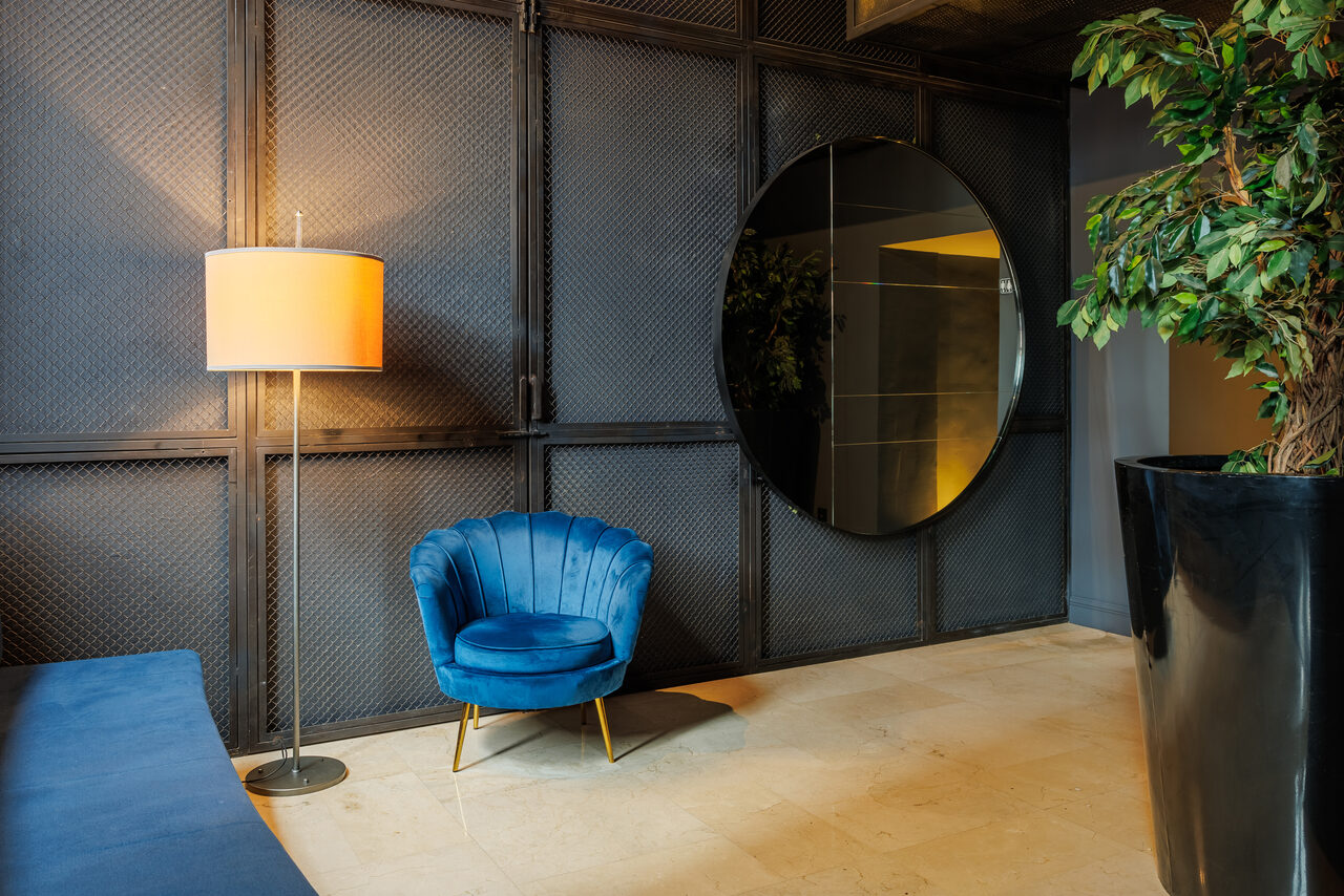 Арендовать Zafferano Loft(Зафферано лофт). Комната с лампой на высокой ножке с тканевым абажуром, синим креслом на металлических ножках и круглым зеркалом на стене. Стена с темной текстурированной поверхностью, светлый пол. В углу крупное растение в черном горшке.