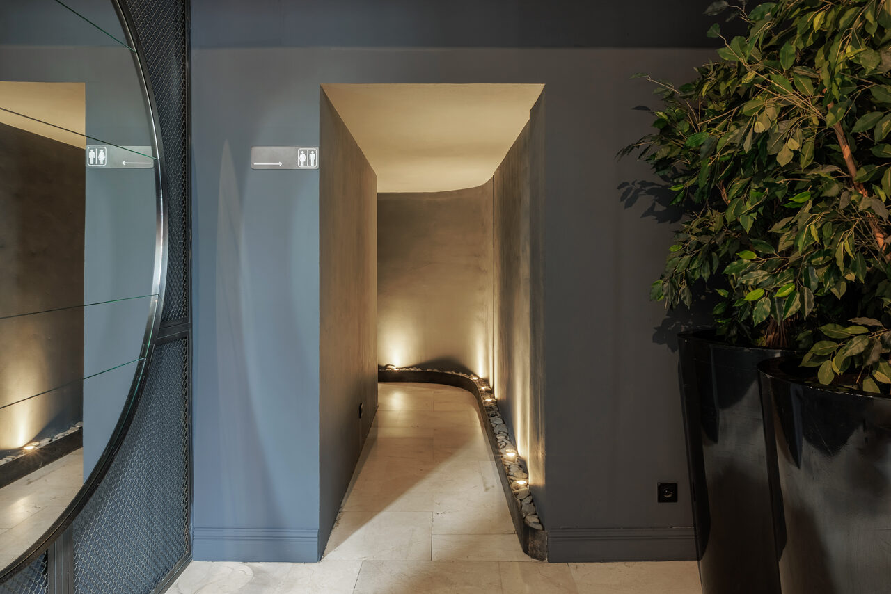 Арендовать Zafferano Loft(Зафферано лофт). Коридор с серыми стенами и светлым полом, в стене ниша с подсветкой. Справа темная большая кадка с растением. Слева часть круглого зеркала на стене. Освещение точечное, встроенное. Декор современный.