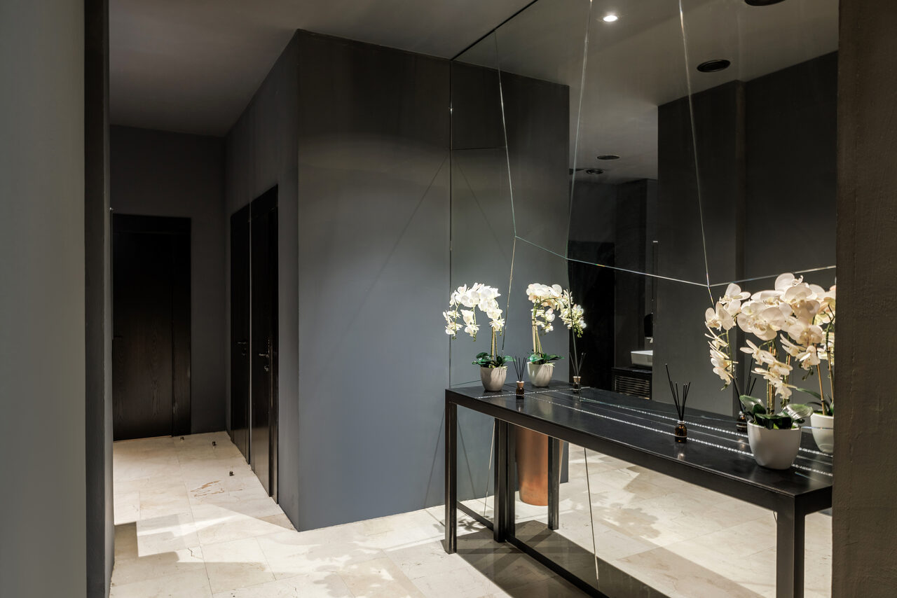 Арендовать Zafferano Loft(Зафферано лофт). Коридор с тёмно-серыми стенами. Зеркальная стена с правой стороны. Белый пол. Чёрный стол с белыми цветами в горшках и украшениями. Точечное освещение на потолке. Тёмные дверные проёмы.