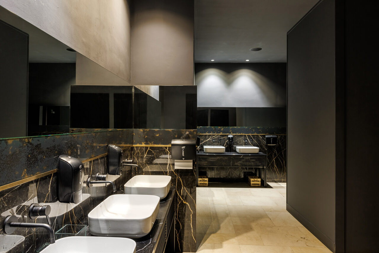 Арендовать Zafferano Loft(Зафферано лофт). Современная туалетная комната с двумя умывальниками из белой керамики на черной мраморной поверхности. Черные автоматические диспенсеры для мыла. Зеркала на стене. Бежевый каменный пол. Мягкое потолочное освещение. Тёмные стены и перегородки.