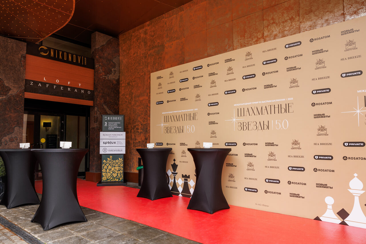 Арендовать Zafferano Loft(Зафферано лофт). Вход в здание с вывеской, покрытый красным ковром. Справа стена с логотипами и текстом "Шахматные звезды 150". Три высоких круглых столика в черных скатертях. На заднем плане видна дверь с надписью "LOFT ZAFERANO".