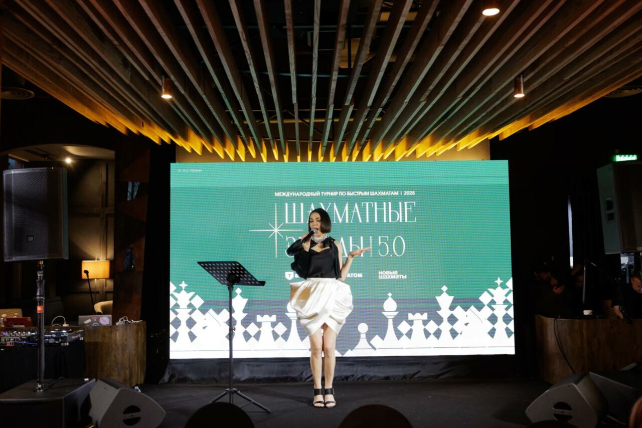 Арендовать Zafferano Loft(Зафферано лофт). Просторная сцена с деревянным потолком, освещение точечными светильниками, на сцене микрофонная стойка, крупный экран с зеленым фоном и белым текстом. По бокам колонки, сбоку видна мебель и техника. Макет шахматных фигур на экране.