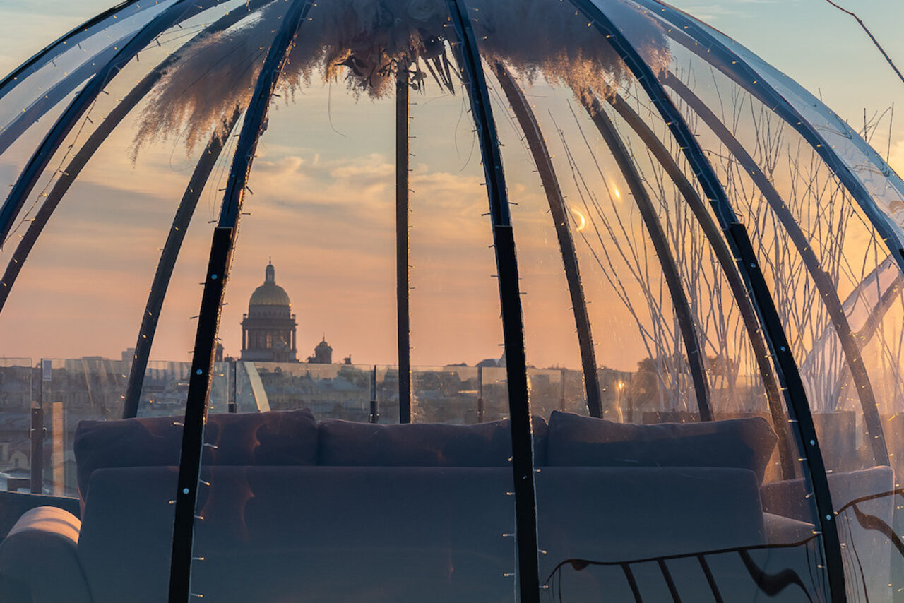 Арендовать Sky Terrasse(Небо Терраса). Прозрачный купол с панорамным видом на город, конструкция из металлических дуг. Внутри темный диван, светодиодная подсветка, подвесной декор. На горизонте городской пейзаж с куполом большого здания, закатное освещение.