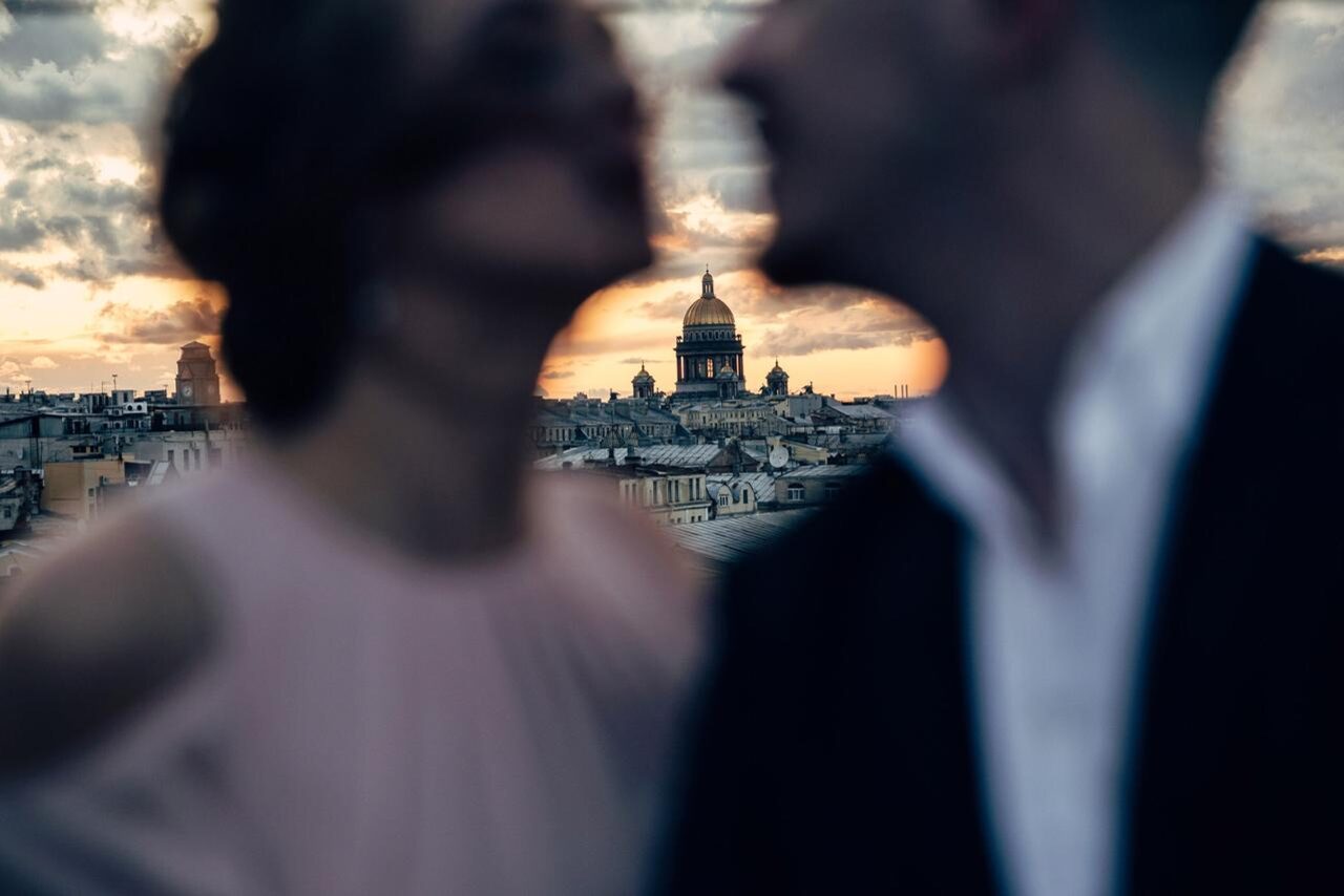 Арендовать Sky Terrasse(Небо Терраса). Изображены размытые силуэты двух человек на фоне городской панорамы с куполом, при закатном освещении. Видны крыши зданий, облачное небо с оранжевыми и фиолетовыми оттенками.